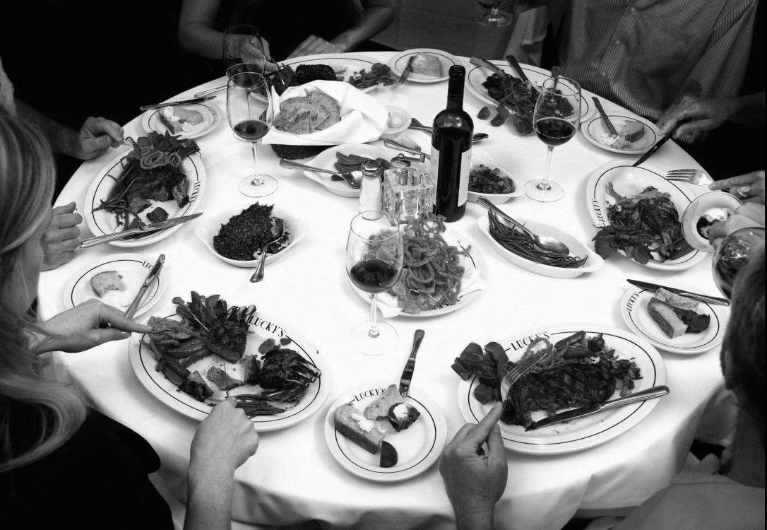 Overhead bustling view of a large dining table covered with premium steak dishes, fresh sides, and craft cocktails